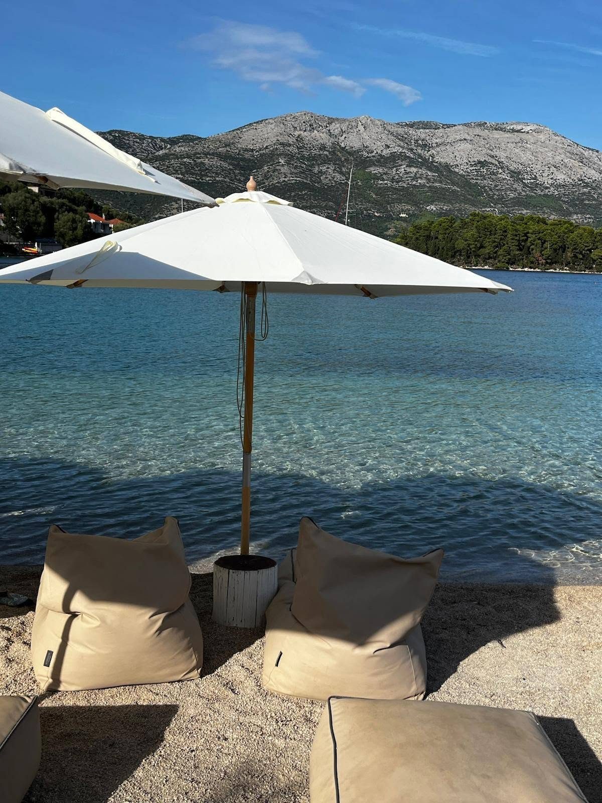 A white parasol and two cushions on a pebble beach, clear sea and mountains behind