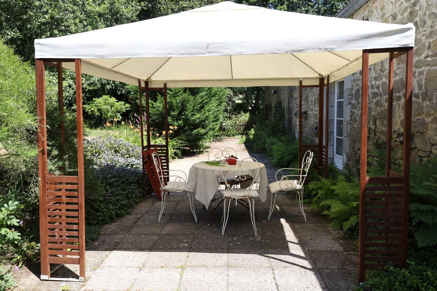 A table and white iron chairs under a garden canopy beside the stone house